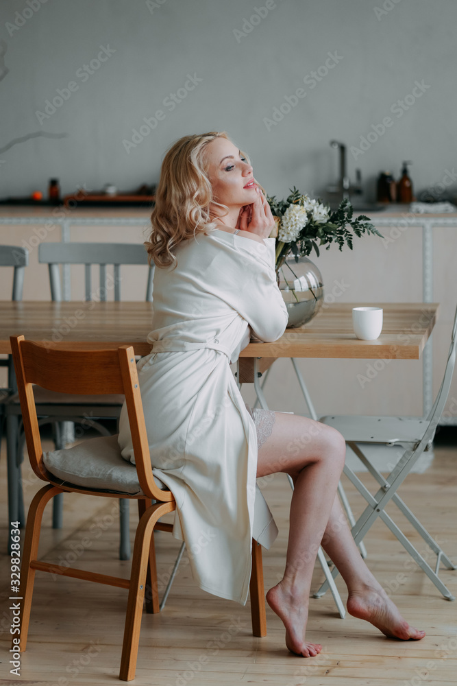 Bride's bouquet. Beautiful happy happy girl blonde bride in a white light sexy robe in the morning in the kitchen is happy, smiling and touching a bouquet of fresh flowers , preparing for the wedding Bride's bouquet. Beautiful happy happy girl blonde bride in a white light sexy robe in the morning in the kitchen is happy, smiling and touching a bouquet of fresh flowers , preparing for the wedding