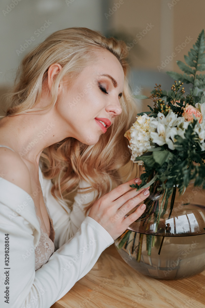 Naklejka premium Bride's bouquet. Beautiful happy happy girl blonde bride in a white light sexy robe in the morning in the kitchen is happy, smiling and touching a bouquet of fresh flowers , preparing for the wedding