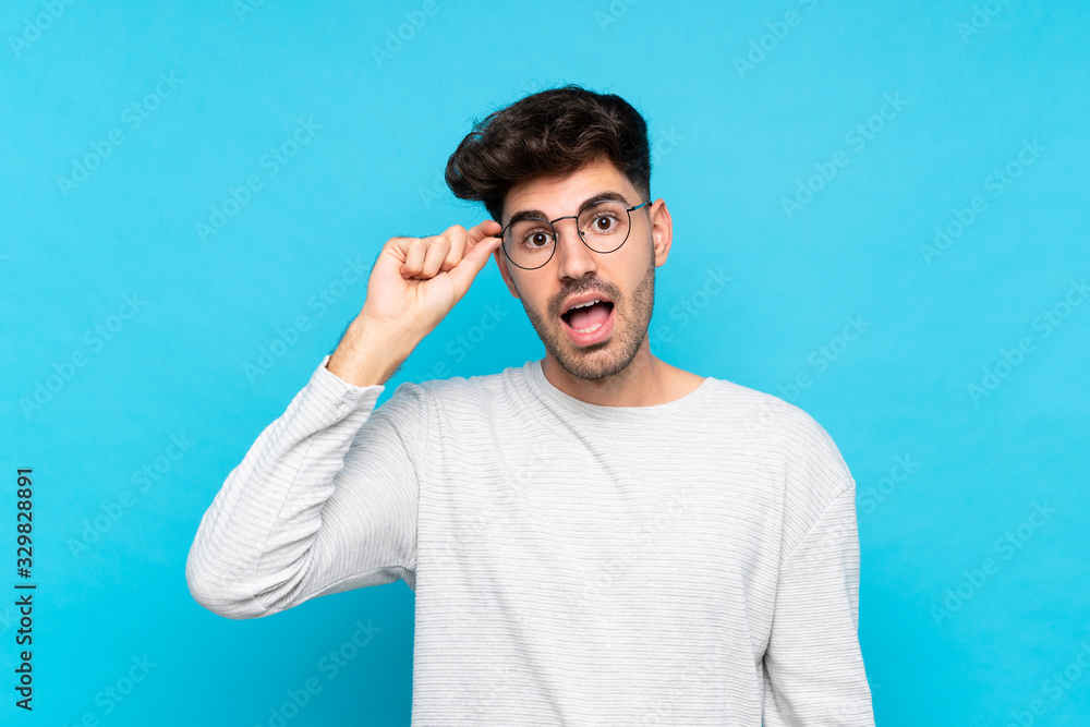 Young man over isolated blue background with glasses and surprised Young man over isolated blue background with glasses and surprised