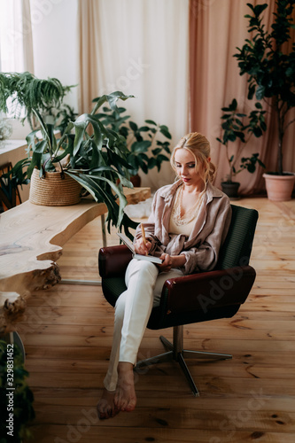 A thoughtful serious girl blogger freelancer works at home on a laptop, writes ideas in her notebook diary, and organizes in a bright spacious office with a view of the city decorated with greenery
