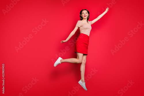 Full length profile photo of pretty lady jump high rejoicing summer warm weather flying up to sky imagination wear casual striped t-shirt red skirt isolated red bright color background