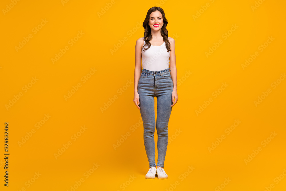 Full body photo of funny pretty shiny lady standing one place good mood slim figure shapes wear white tank-top jeans footwear isolated bright yellow color background Full body photo of funny pretty shiny lady standing one place good mood slim figure shapes wear white tank-top jeans footwear isolated bright yellow color background