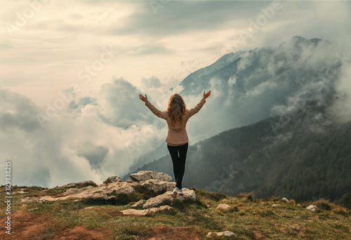 Tourist Woman standing on top mountain turned away raised arms enjoy divine view mountains in white clouds. Mysterious silhouette. Backdrop sunshine sky green forest in fog. Peak Italy Monte Baldo
