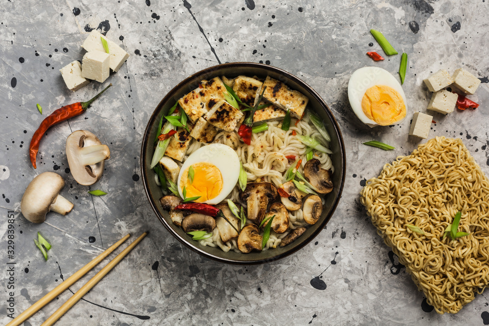 Noodle bowl with mushrooms, egg, tofu cheese, chopsticks and ingredients on a gray background. Asian food.
