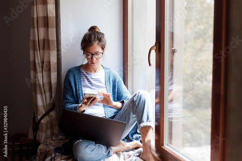 Young woman working from home office. Freelancer using laptop, phone and the Internet. Workplace in living room on windowsill. Concept of female business, career, shopping online. Lifestyle moment. photo