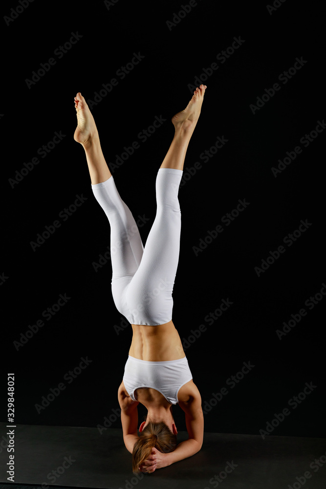 young woman in white sportswear doing yoga handstand exercise isolated on black