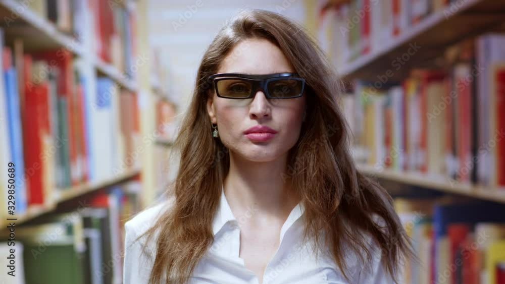 Young brunette woman working in the library wearing black virtual reality glasses using technology in modern education. Background of bookshelves. Young brunette woman working in the library wearing black virtual reality glasses using technology in modern education. Background of bookshelves.