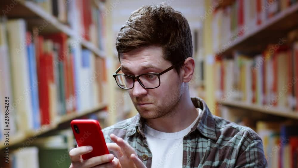 Smart appealing young man wears glasses reading news on smartphone rejoicing shocked facial expression standing near bookshelves. In the library.