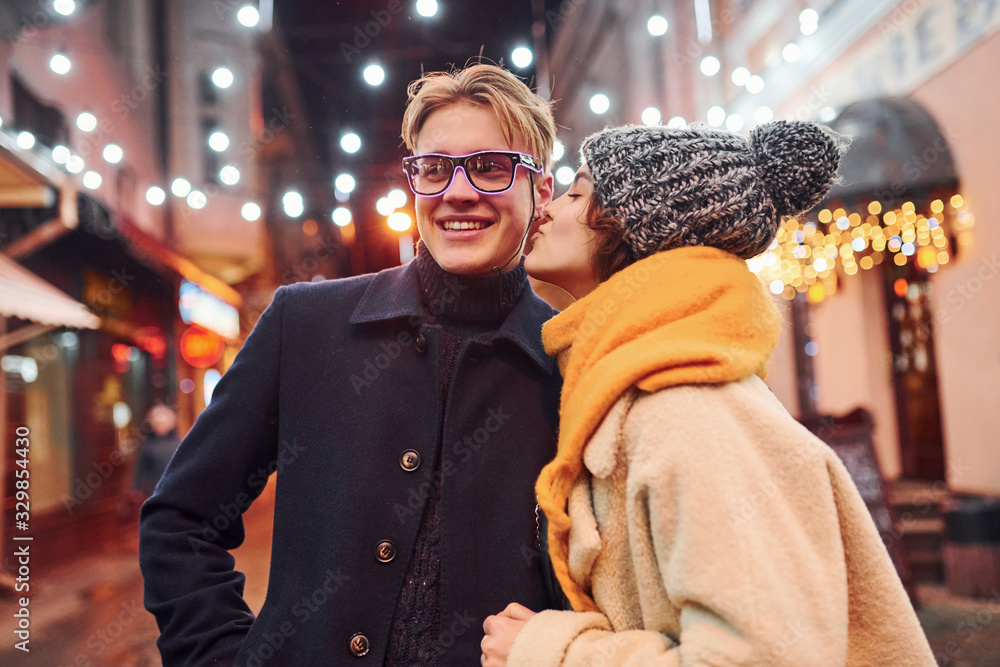 Young girl standing on christmas decorated street and kisses her boyfriend