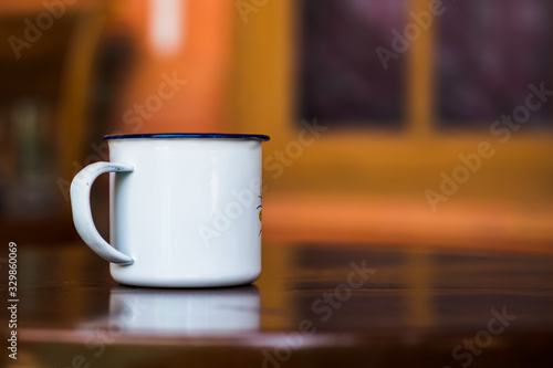 Vintage enamel mug on a classic teak table against simple window background in blur. Antique rustic coffee cup on a brown  smooth wooden surface.