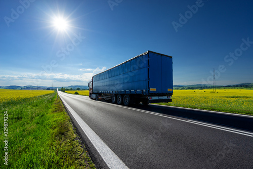 Truck driving on the asphalt road between the yellow flowering rapeseed fields under radiant sun in the rural landscape