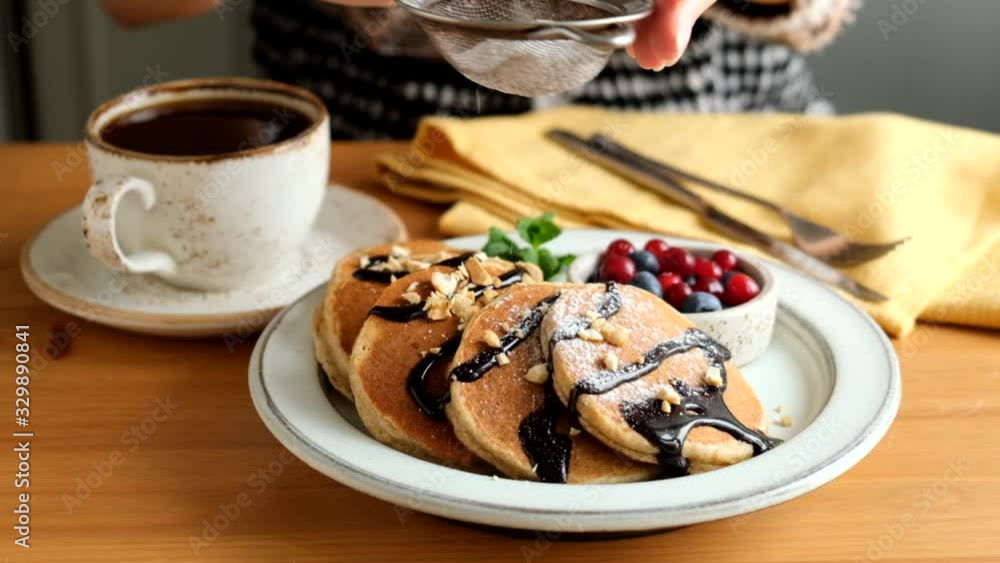 Slow motion of cook sift confectioner sugar on pancakes served with chocolate sauce, berries and cup of black coffee. Tasty sweet breakfast on a wooden table Slow motion of cook sift confectioner sugar on pancakes served with chocolate sauce, berries and cup of black coffee. Tasty sweet breakfast on a wooden table