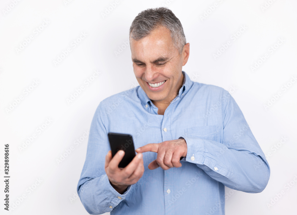 adult handsome smiling man with grey hair wearing blue shirt pointing at his smartphone laughing ,standing on isolated white background, modern technology concept adult handsome smiling man with grey hair wearing blue shirt pointing at his smartphone laughing ,standing on isolated white background, modern technology concept
