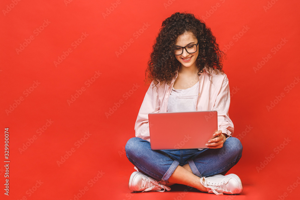 Happy young curly beautiful woman sitting on the floor with crossed legs and using laptop on red background. Happy young curly beautiful woman sitting on the floor with crossed legs and using laptop on red background.