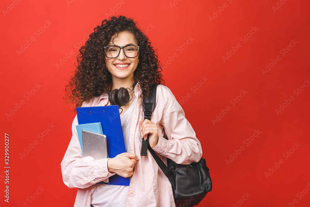 Young curly student woman wearing backpack glasses holding books and tablet over isolated red background. - obrazy, fototapety, plakaty 