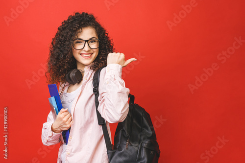 Fotografie, Obraz Young curly student woman wearing backpack glasses holding books and tablet over isolated red background