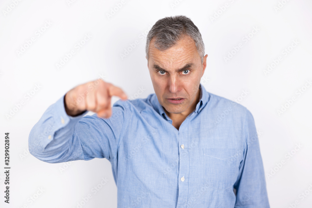 adult angry man with grey hair wearing blue shirt pointing at somebody looks furious standing on isolated white background, body language concept adult angry man with grey hair wearing blue shirt pointing at somebody looks furious standing on isolated white background, body language concept