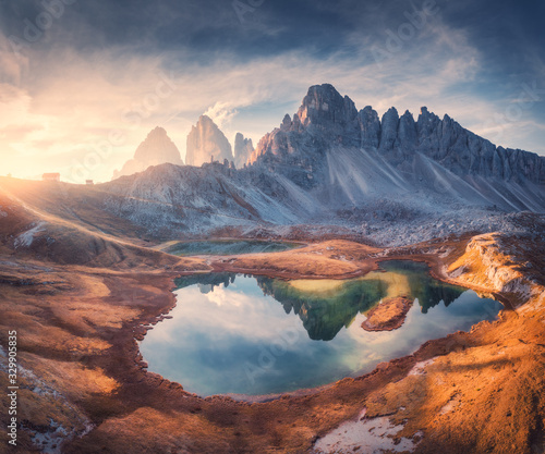 Aerial view of beautiful rocks  mountain lake  reflection in water and houses on the hill at sunset. Autumn landscape with mountains  blue sky and sunlight. Dolomites  Italy. Top view of Italian alps