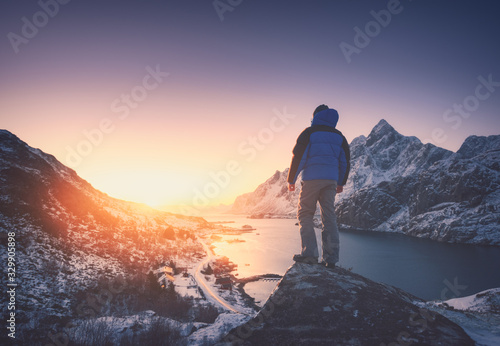 Man is standing on the mountain peak against snowy mountains, fjord at colorful sunset in winter. Young man on the stone, sea coast and rocks, blue sky in Lofoten Islands, Norway. Travel and hiking