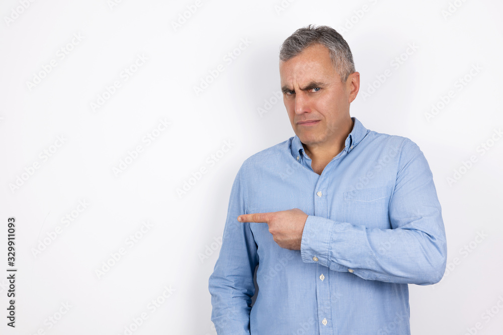 mature handsome confused man with grey hair wearing blue shirt standing on isolated white background points with his fingers looking bewildered, body language concept mature handsome confused man with grey hair wearing blue shirt standing on isolated white background points with his fingers looking bewildered, body language concept