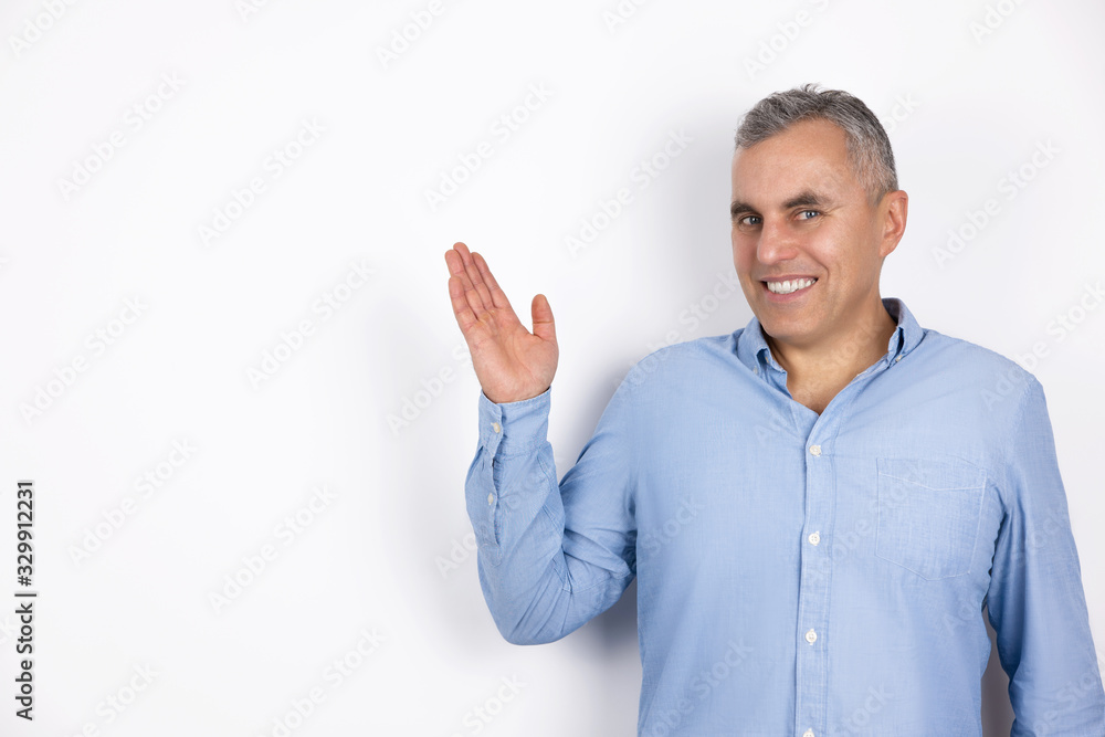 mature smiling man with grey hair wearing blue shirt standing on isolated white background holds his hand up and to the side, body language concept mature smiling man with grey hair wearing blue shirt standing on isolated white background holds his hand up and to the side, body language concept
