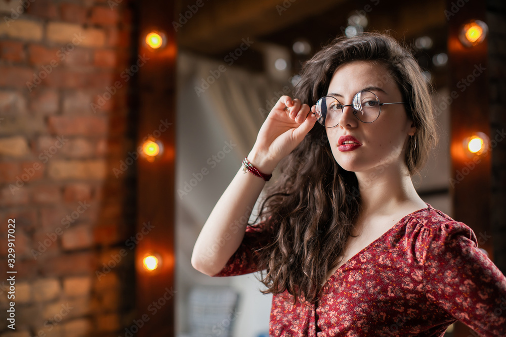 Close up portrait of attractive student girl with curly hair, in round glasses at loft apartment. Young woman looking in camera and ressettling her spectacles.