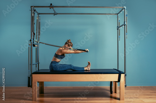 Fototapeta Young girl doing pilates exercises with a reformer bed. Beautiful slim fitness trainer on reformer gray background, low key, art light. Fitness concept