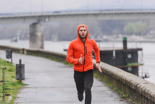 Handsome young athlete man running fast along river in orange windbreaker jacket. Extreame weather sport. Running on a rainy day. photo