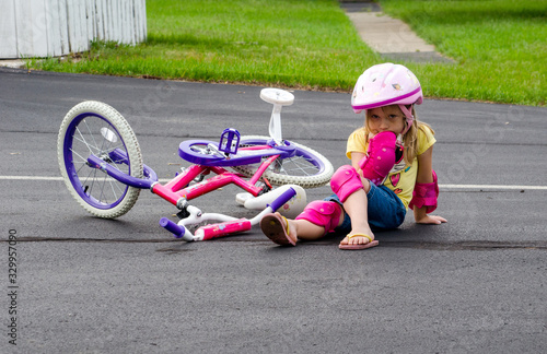 A little girl in safety gear, sits in the middle of a drive after falling off her new bike.