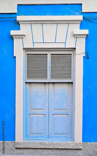 Tall classic ornamental closed window with shutters in a blue wall.