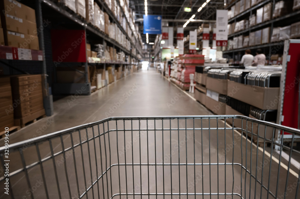 Fototapeta premium Empty shopping cart in a furniture store warehouse. Supermarket shopping basket aisle with box shelf abstract blur defocused background