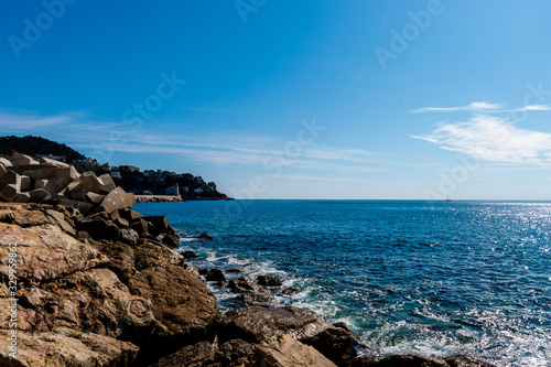 The Mediterranean Sea waves reaching the big rocks on the shore with the sunlight reflecting in the turquoise water and a peninsula in the background on a sunny day  Nice  France 