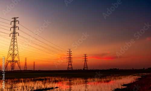 High voltage electric pylon and electrical wire with sunset sky. Electricity poles. Power and energy concept. High voltage grid tower with wire cable at rice farm field near industrial factory.