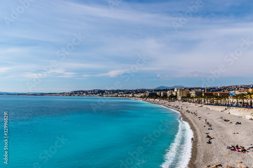 Panoramic wide angle shot of the Quai des États-Unis, people relaxing on the beach of the Mediterranean Sea coastline with the sunlight reflecting in the turquoise water (Nice, France)