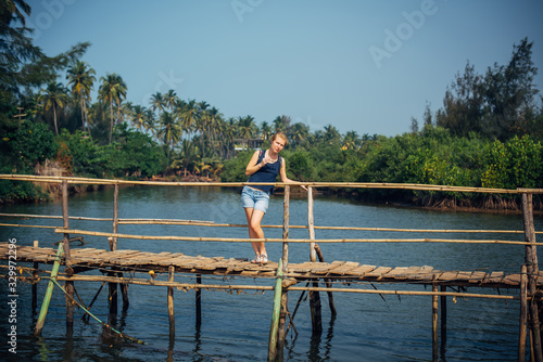 A female tourist in denim shorts with short haircut on sunny tropical day. Young pretty girl stands on wooden bridge over small river against palm jungle and blue sky. Trips to picturesque places.