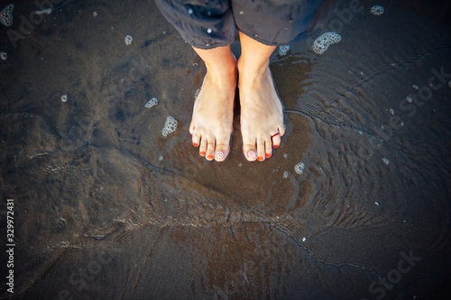 Woman's feet on sandy beach, standing in ocean water, copy space. Pedicured feet with red nail polish on toes in the sea. Bare feet standing on sand, top view. Hello summer, welcome vacation concept.