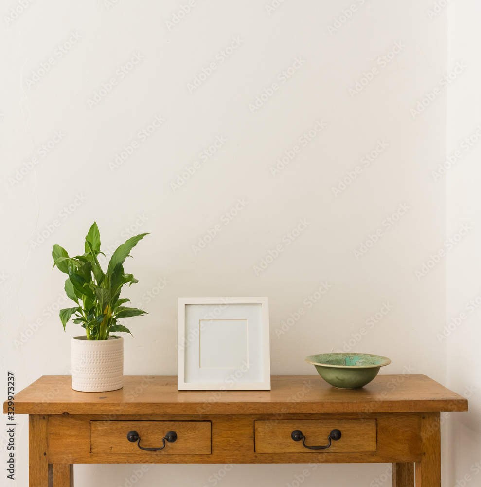 Green leafy houseplant in white pot with blank square picture frame and ceramic bowl on oak side table against white wall Green leafy houseplant in white pot with blank square picture frame and ceramic bowl on oak side table against white wall