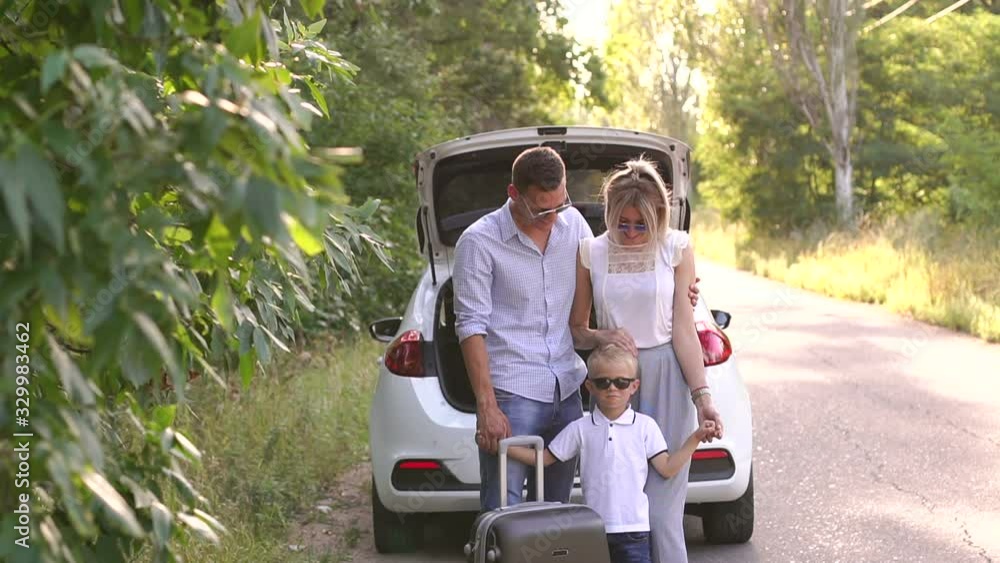 Portrait of a happy young family with a suitcase near the trunk of a car on an empty road in the woods. The family travels by car. Parents with son gather on summer holidays. Portrait of a happy young family with a suitcase near the trunk of a car on an empty road in the woods. The family travels by car. Parents with son gather on summer holidays.