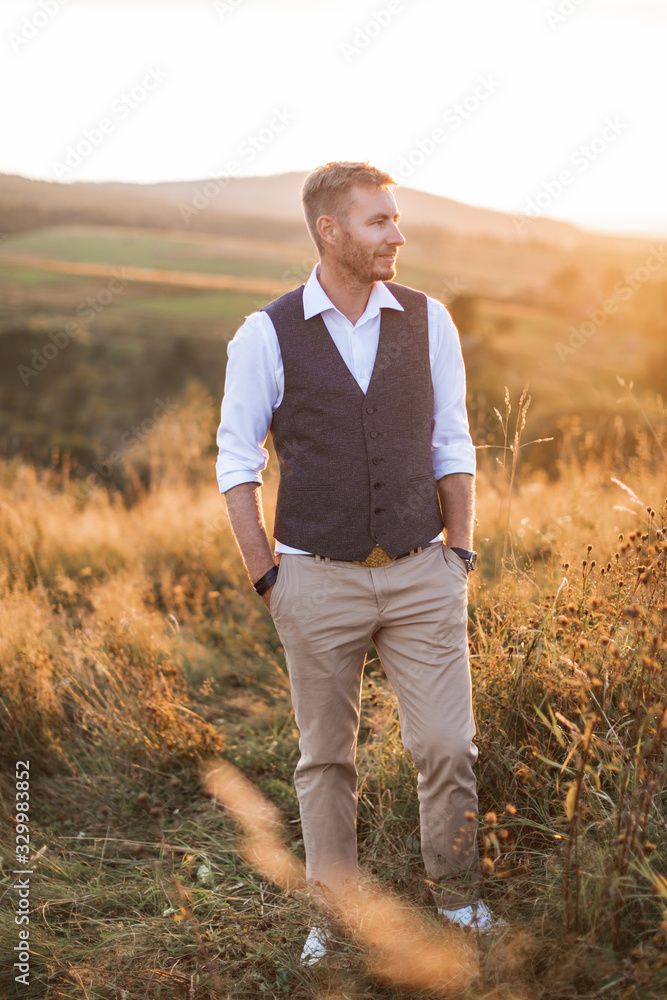Portrait of handsome young man, farmer or cowboy, wearing casual suit, looking to a side, while standing in summer field in countryside, sunset