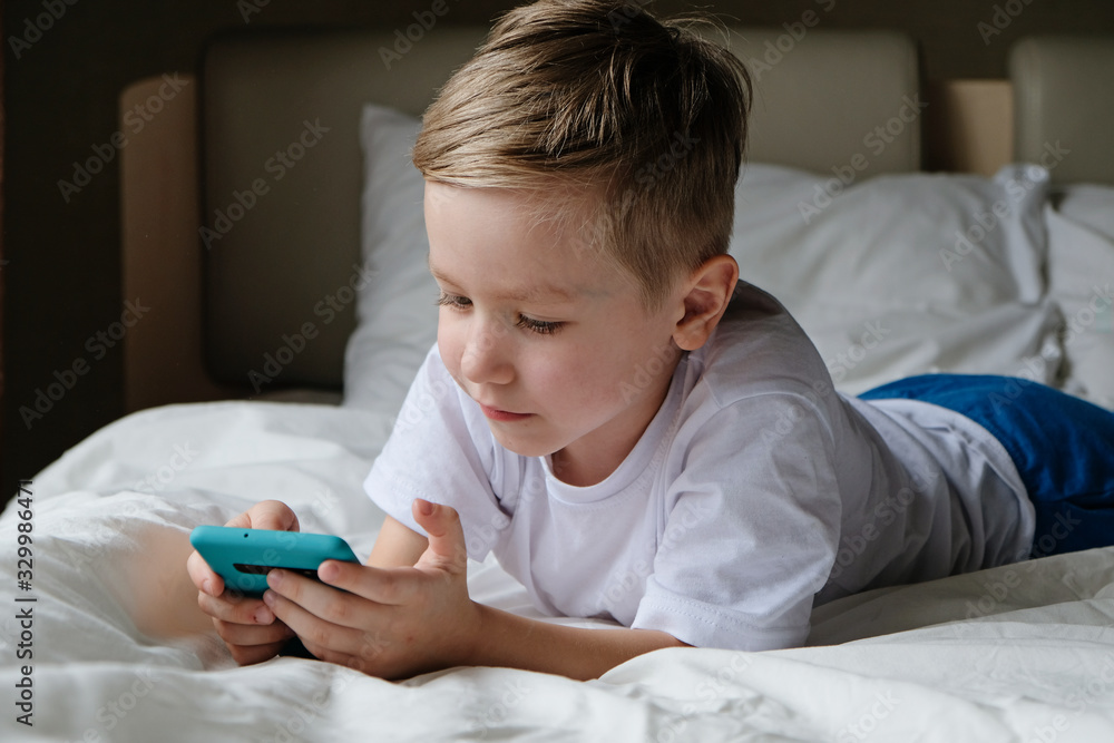 Naklejka premium Cute little boy toddler playing mobile game, lying on a bed and holding smartphone