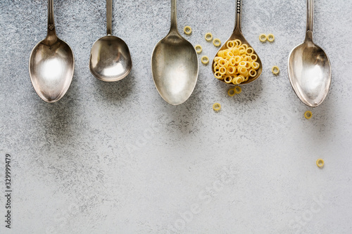 Anellini pasta in spoon and empty iron spoons on gray concrete background. Cooking concept. Top view.