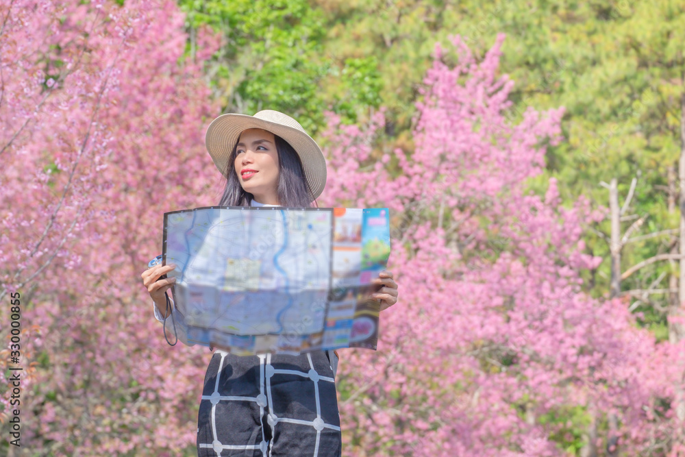 Naklejka premium Smiling woman traveler in pink sakura blossom park at doi inthanon landmark chiang mai thailand with backpack holding world map on holiday, relaxation concept, travel concept