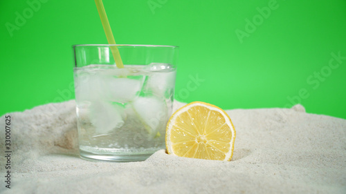 glass with ice and soda water and lemon on the sand on a green background