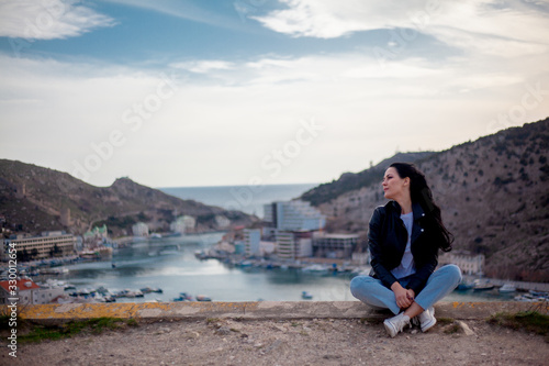 Fashion style woman sit on the top and look at city. The traveler looking to the cityscape panorama by the sea. Girl succesfully travels
