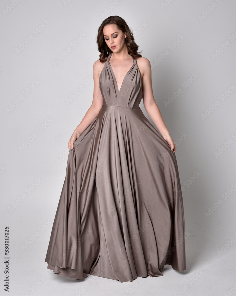 Portrait of a pretty brunette girl wearing a long silver evening gown, full length standing pose against a studio background. Portrait of a pretty brunette girl wearing a long silver evening gown, full length standing pose against a studio background.