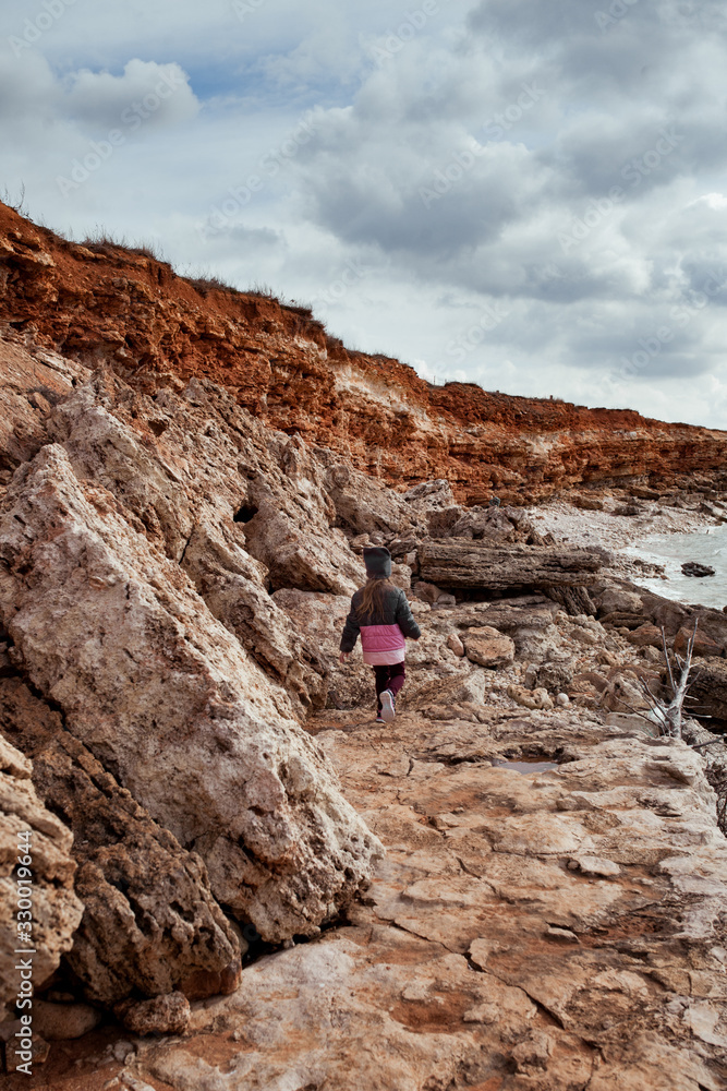 Small girl walks on the sea coast, she is wearing a coat, the kid travels along the sea, outdoor