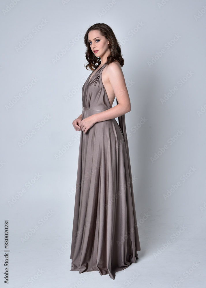 Portrait of a pretty brunette girl wearing a long silver evening gown, full length standing pose against a studio background. Portrait of a pretty brunette girl wearing a long silver evening gown, full length standing pose against a studio background.