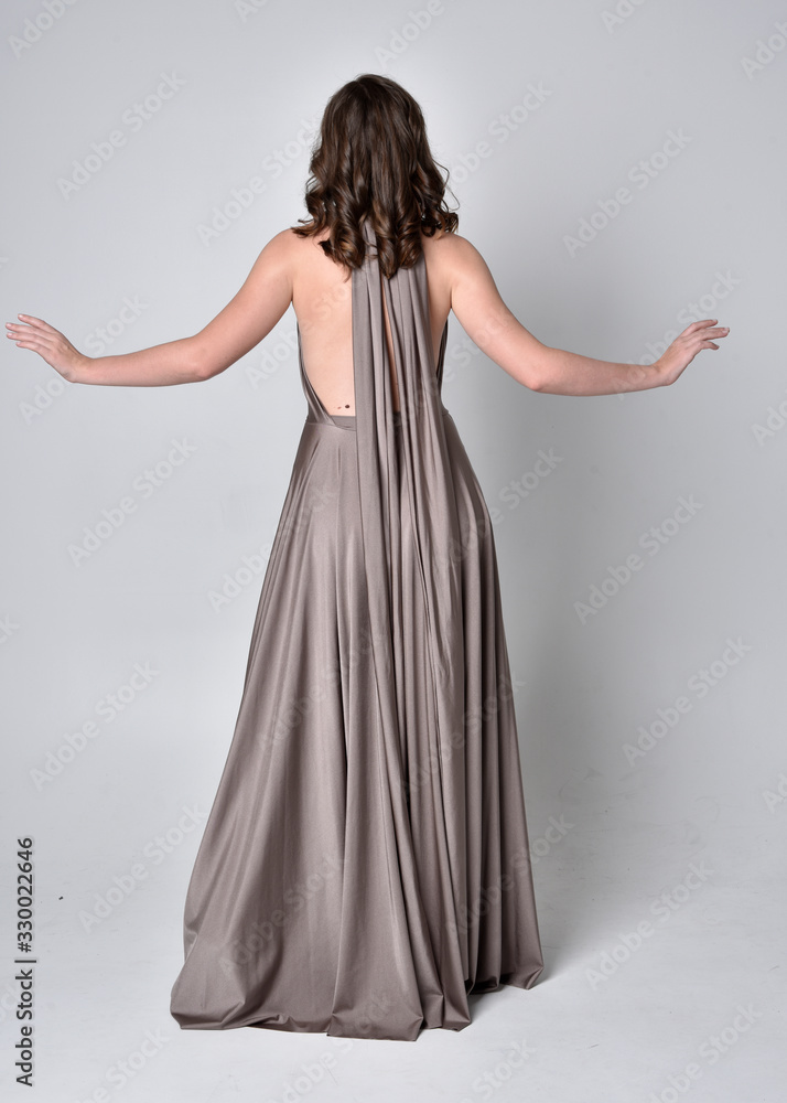 Portrait of a pretty brunette girl wearing a long silver evening gown, full length standing pose against a studio background. Portrait of a pretty brunette girl wearing a long silver evening gown, full length standing pose against a studio background.