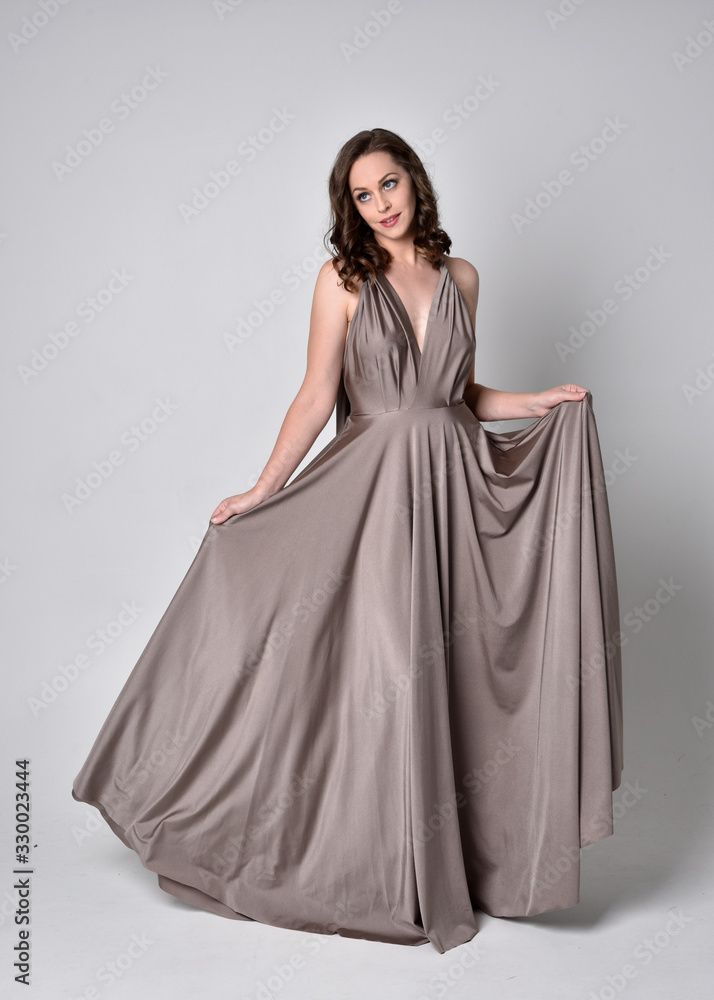  Portrait of a pretty brunette girl wearing a long silver evening gown, full length standing pose against a studio background.