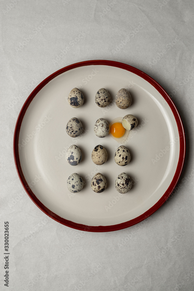 Quail eggs lying in a row on white round plate with red edge. One egg is broken, the yolk leaked. Flat lay. Quail eggs lying in a row on white round plate with red edge. One egg is broken, the yolk leaked. Flat lay.